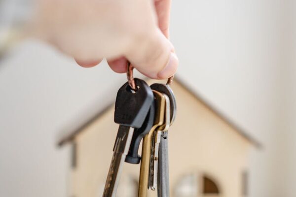 Close-up of a hand holding keys with a miniature wooden house in the background, symbolizing real estate investment.