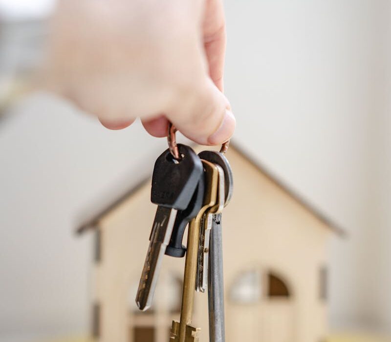 Close-up of a hand holding keys with a miniature wooden house in the background, symbolizing real estate investment.